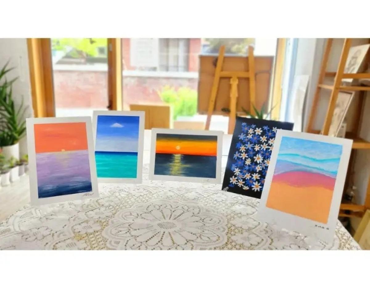 Five small oil pastel landscape and floral paintings on easels displayed on a lace-covered table in a bright studio.