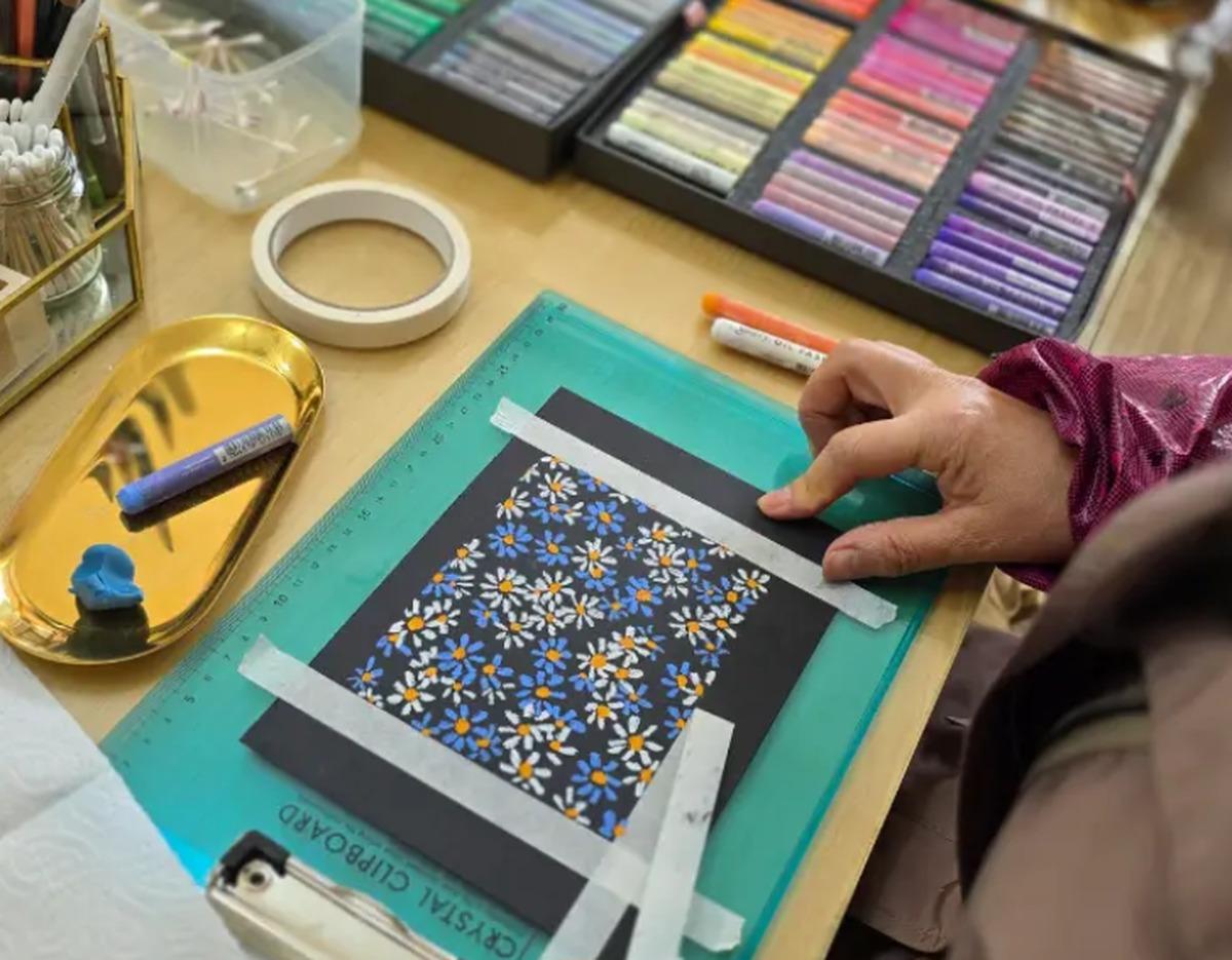 Hand taping black paper on a cutting mat with a completed oil pastel daisy pattern and pastel sticks nearby.