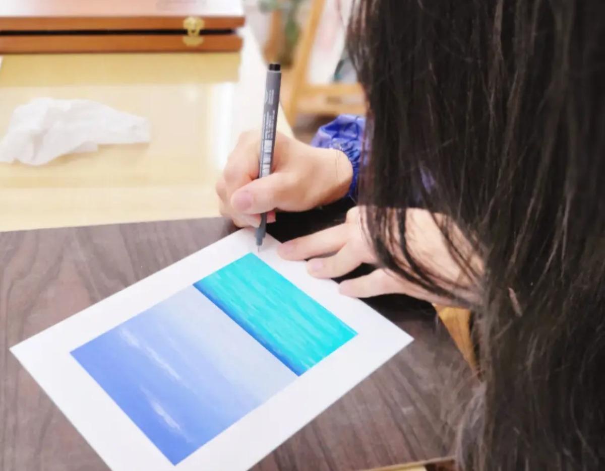 Person with long hair drawing a seascape in blues and turquoise on paper during oil pastel one-hour class in Suwon.