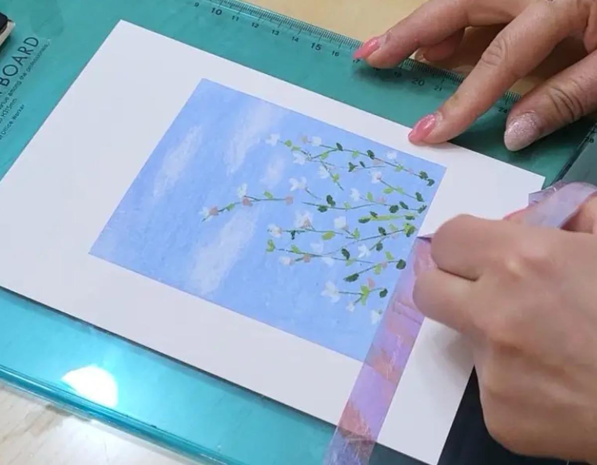 Hands peeling decorative tape from a mounted oil pastel painting of delicate white blossoms on a blue sky background