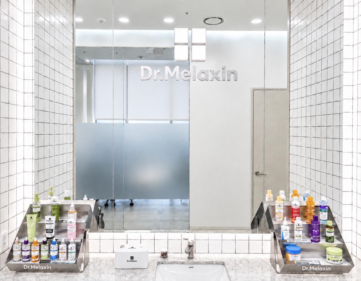 Bright clinic sink and mirror with Dr.Melaxin logo, tiled walls and metal trays of skincare bottles and jars on both sides.