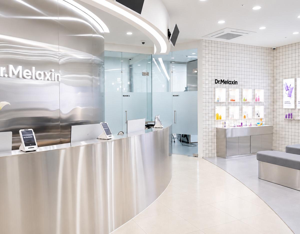 Bright modern clinic reception with curved stainless counter, glass consultation rooms labeled ROOM 1/2, product display wall.