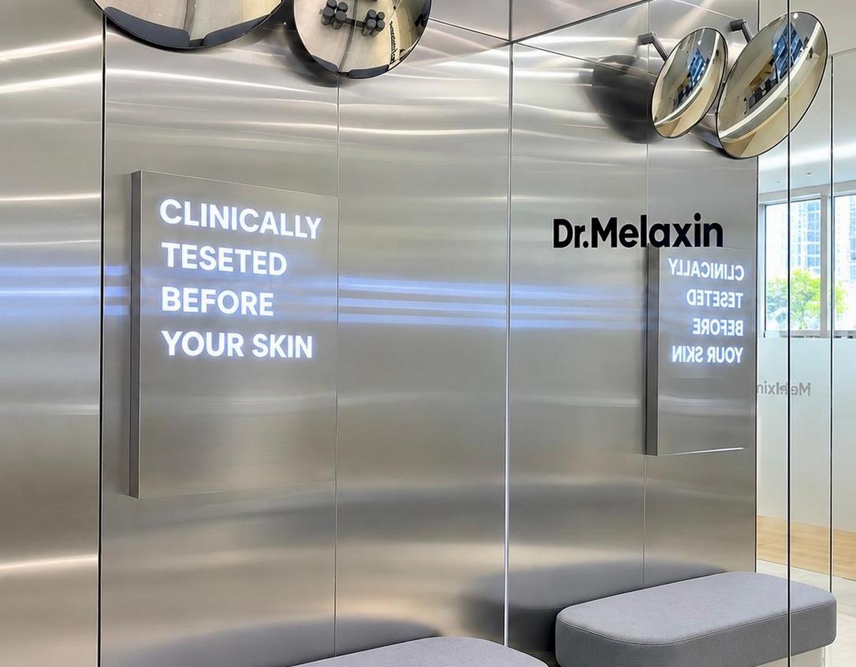 Stainless-steel clinic waiting area with illuminated sign reading "CLINICALLY TESTED BEFORE YOUR SKIN" and gray bench