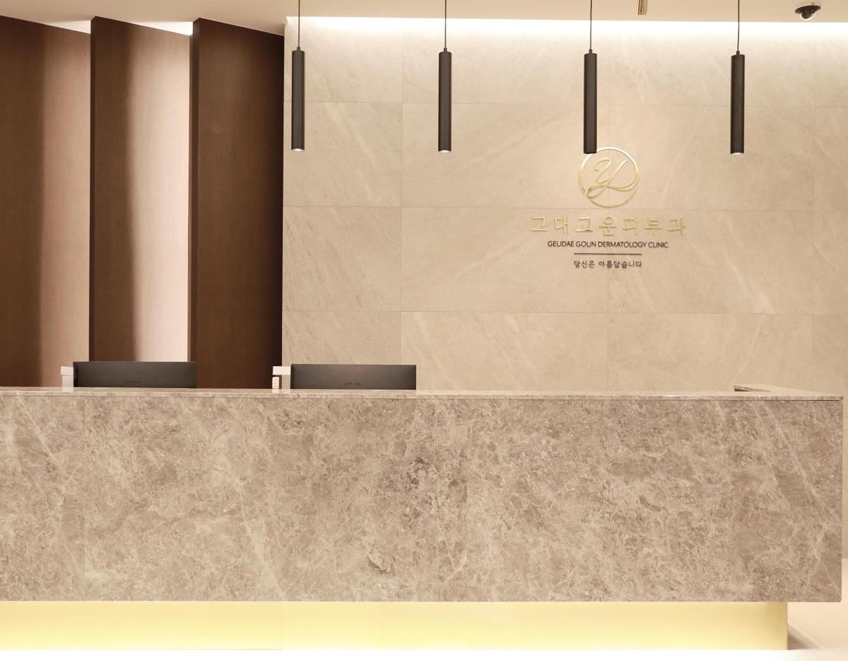 Minimalist marble reception desk with wall signage reading "GEUDAE GOUN DERMATOLOGY CLINIC" and pendant lights above