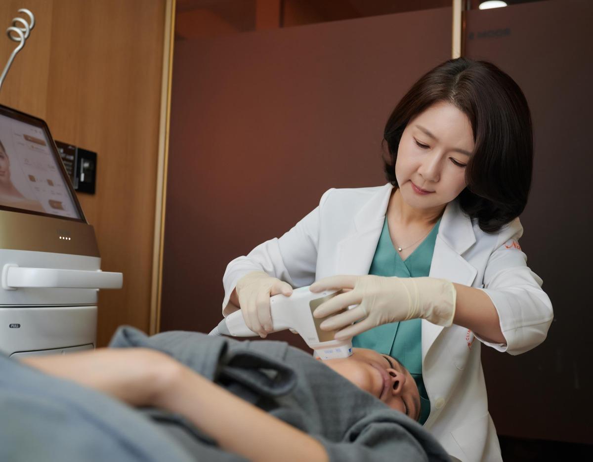 Doctor in white coat performing a handheld ultrasound lifting treatment on a reclining patient's neck in a private clinic room