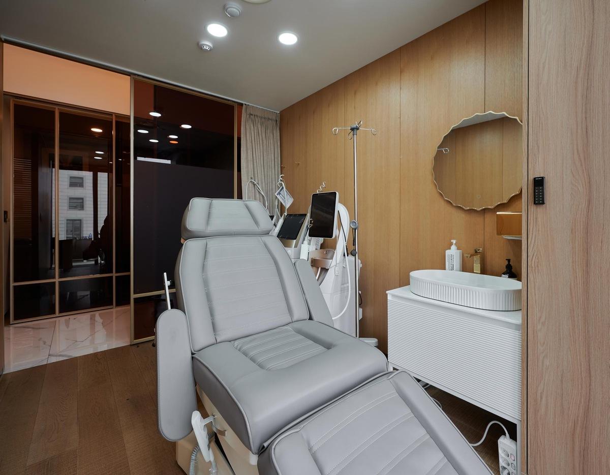 Private treatment room at CHEONGDAM NA CLINIC with gray reclining medical chair, sink, mirror and skincare devices.
