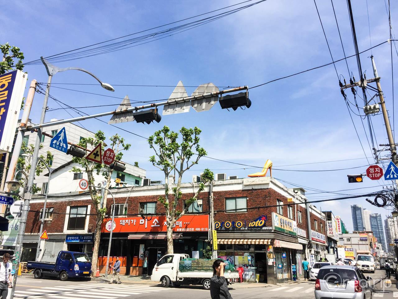 view of the bustling handmade shoe alley in Seongsu-dong, with iconic buildings and a wheel, as cars and electricity run through the thoroughfare under a blue sky.