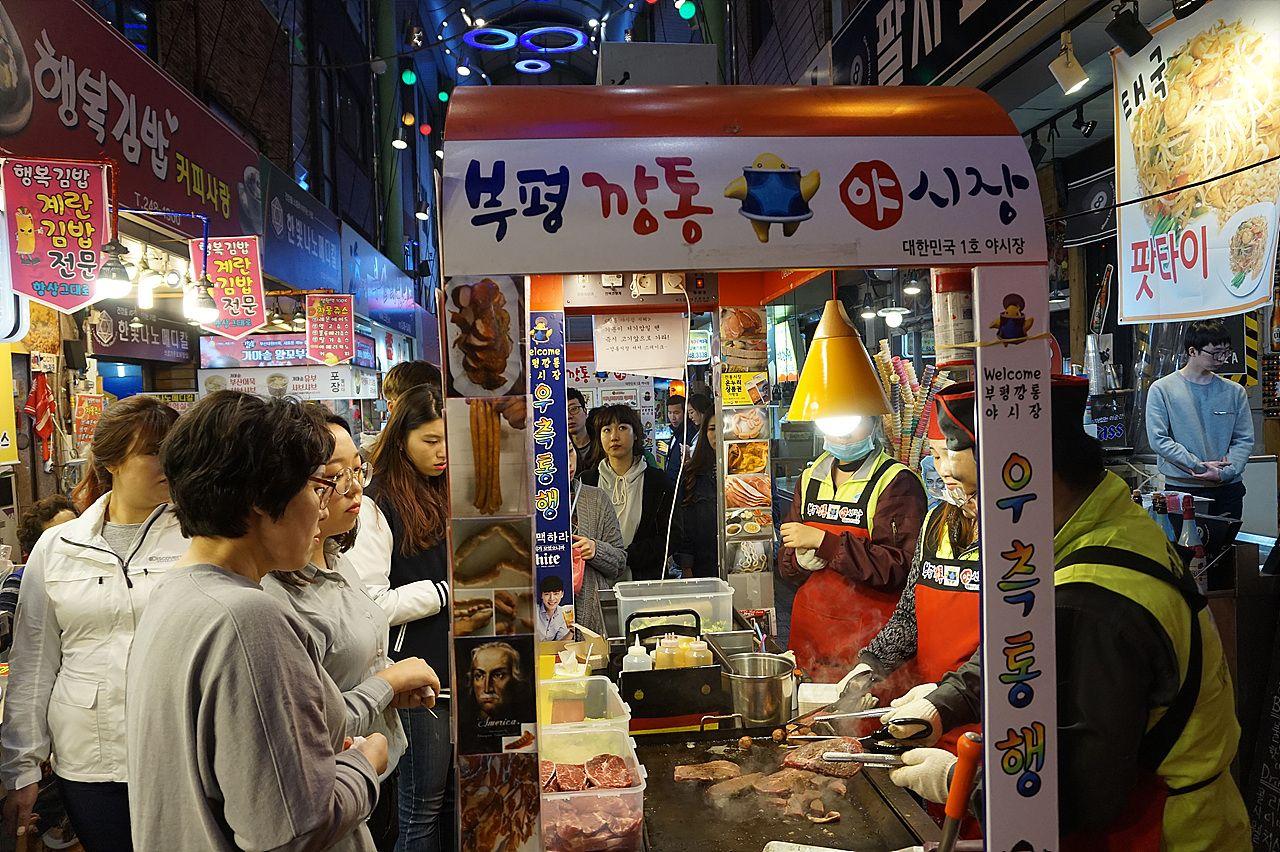 A bustling outdoor market in Korea's 부평깡통시장 district, with a vendor selling outerwear and T-shirts to customers.