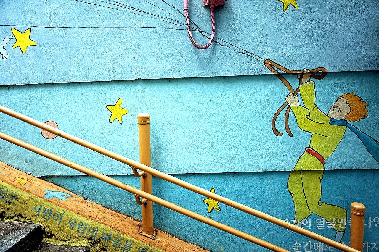 Colorful mural of Little Prince on the wall surrounded by plants in the book alley of Busan. Blue, green, and yellow hues complement the aqua water and rectangular lines.