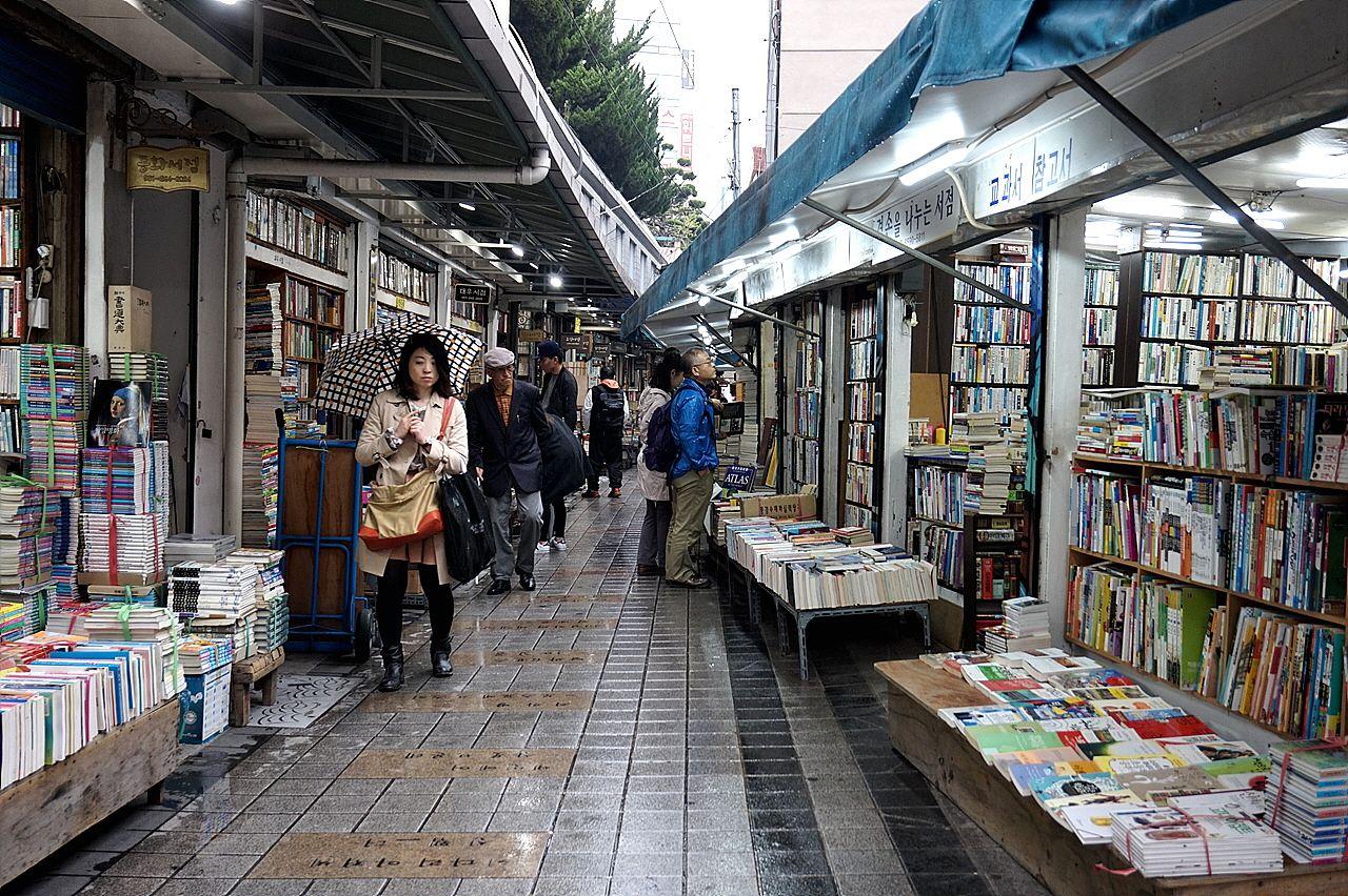 Busy market street in Busan with bookstores, shops and architecture. Customers shopping for publications and retail items, with trade and services available.