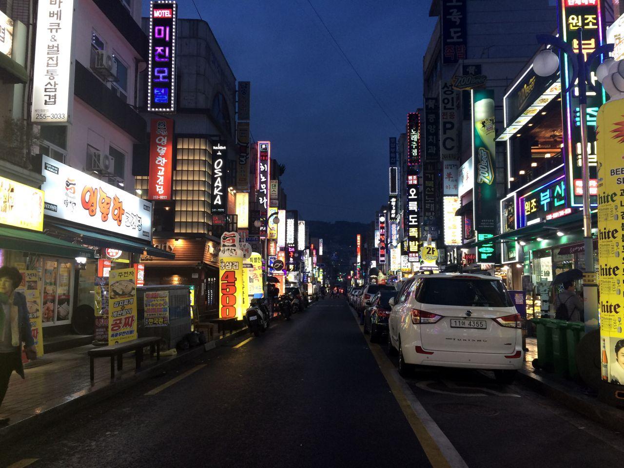 Car parked in front of buildings with vibrant lights, part of the bustling 부평족발골목 area in Korea.