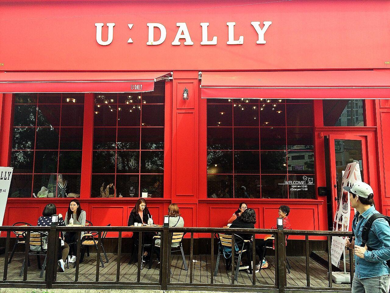 Red chairs and tables arranged outside 카페 유달리, a building with a colorful facade in Seoul, Korea. Guests enjoy leisurely events while bags and luggage line the door and windows.