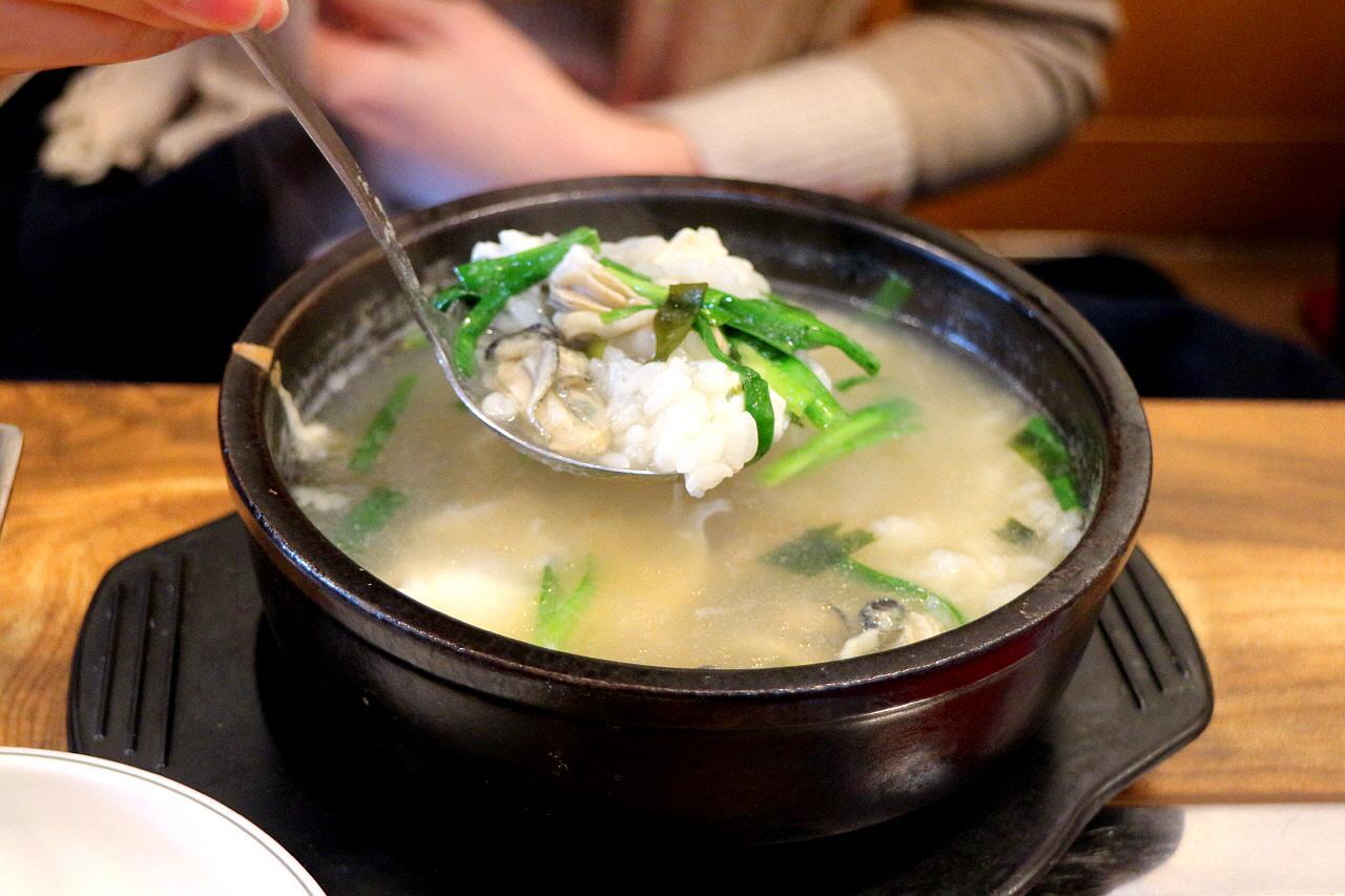 A steaming bowl of 굴국밥 (oyster and rice soup) with ingredients and tableware on the side.