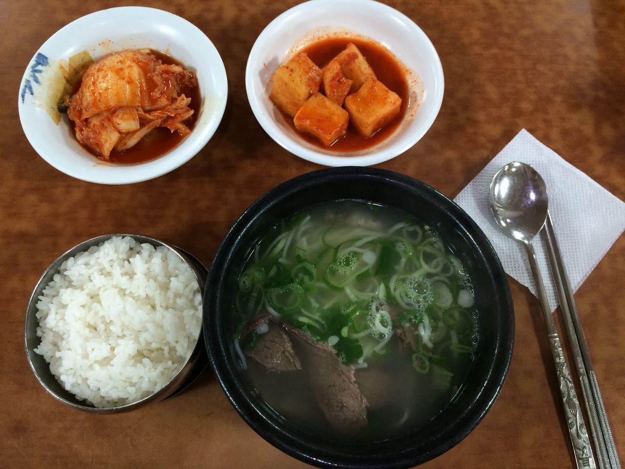 a steaming bowl of 설렁탕, a traditional Korean soup made with sliced beef and clear broth, served with white rice and tableware.