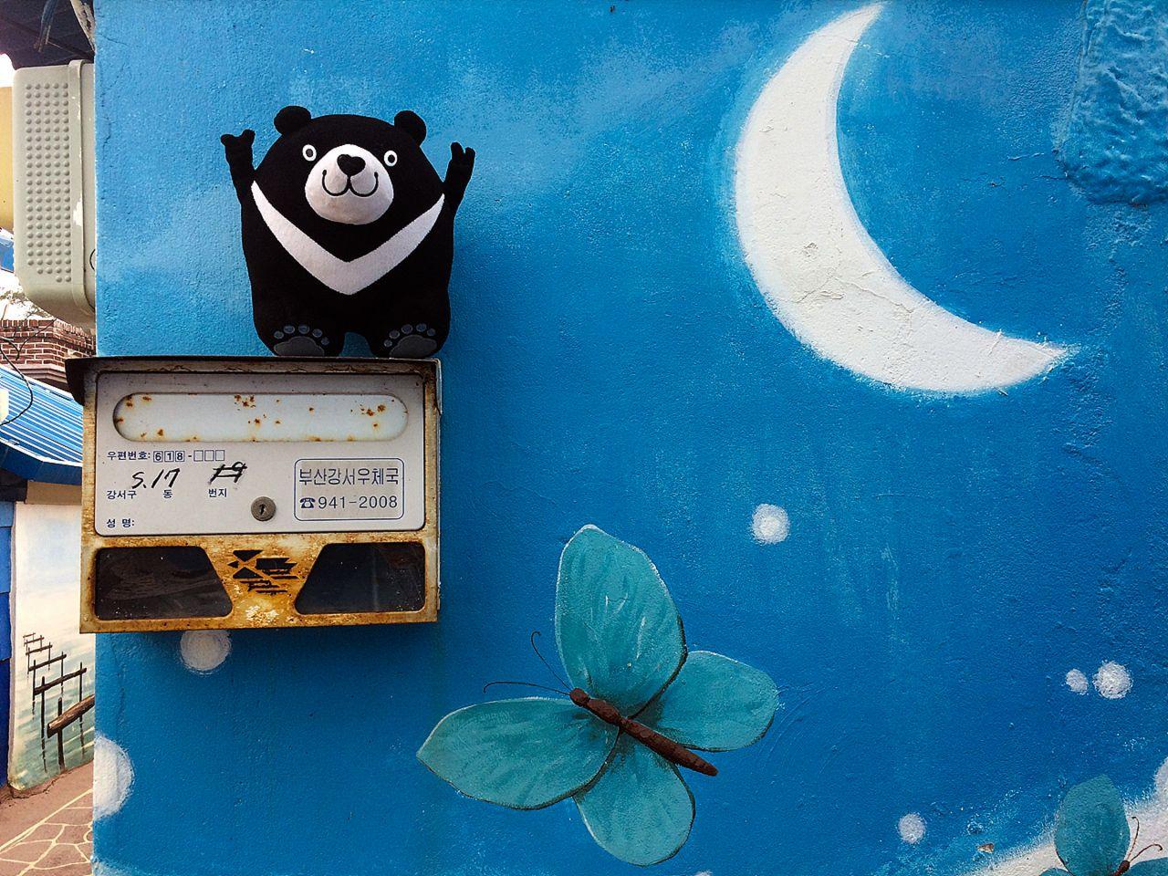 Illustration of a happy village by the azure sea with crescent moon in the sky, inspired by 가덕도 정거마을, 부산, Korea. Artistic font and paint used.