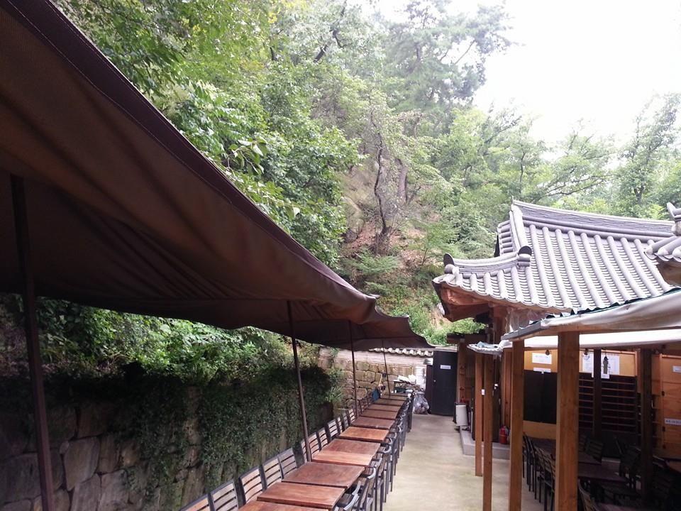 Shaded temple on Namhansanseong Fortress trail with lush botany and wooden roof.