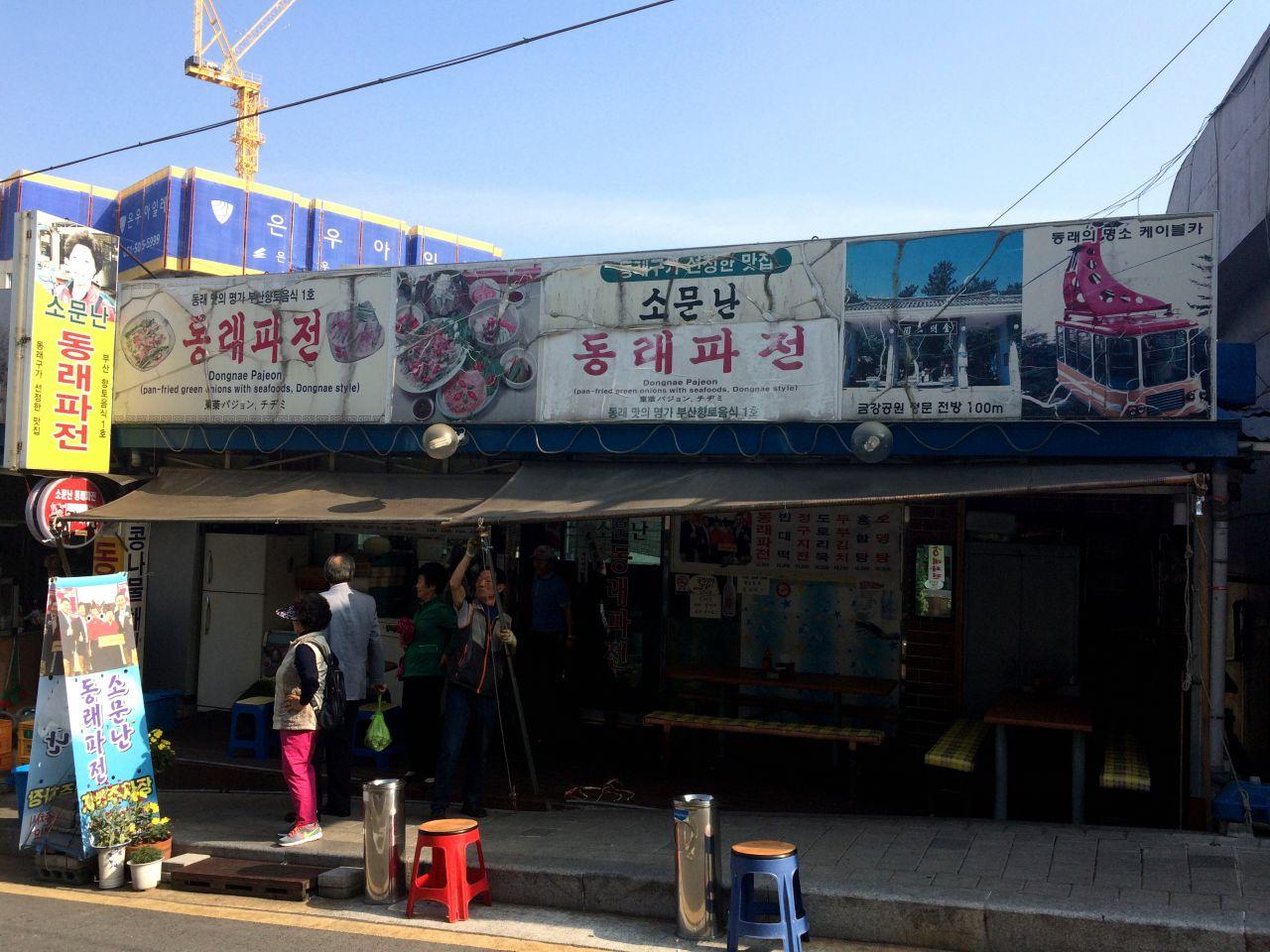 Iconic Dongrae Jeon building in a bustling city, surrounded by markets and art, under a cloudy sky.