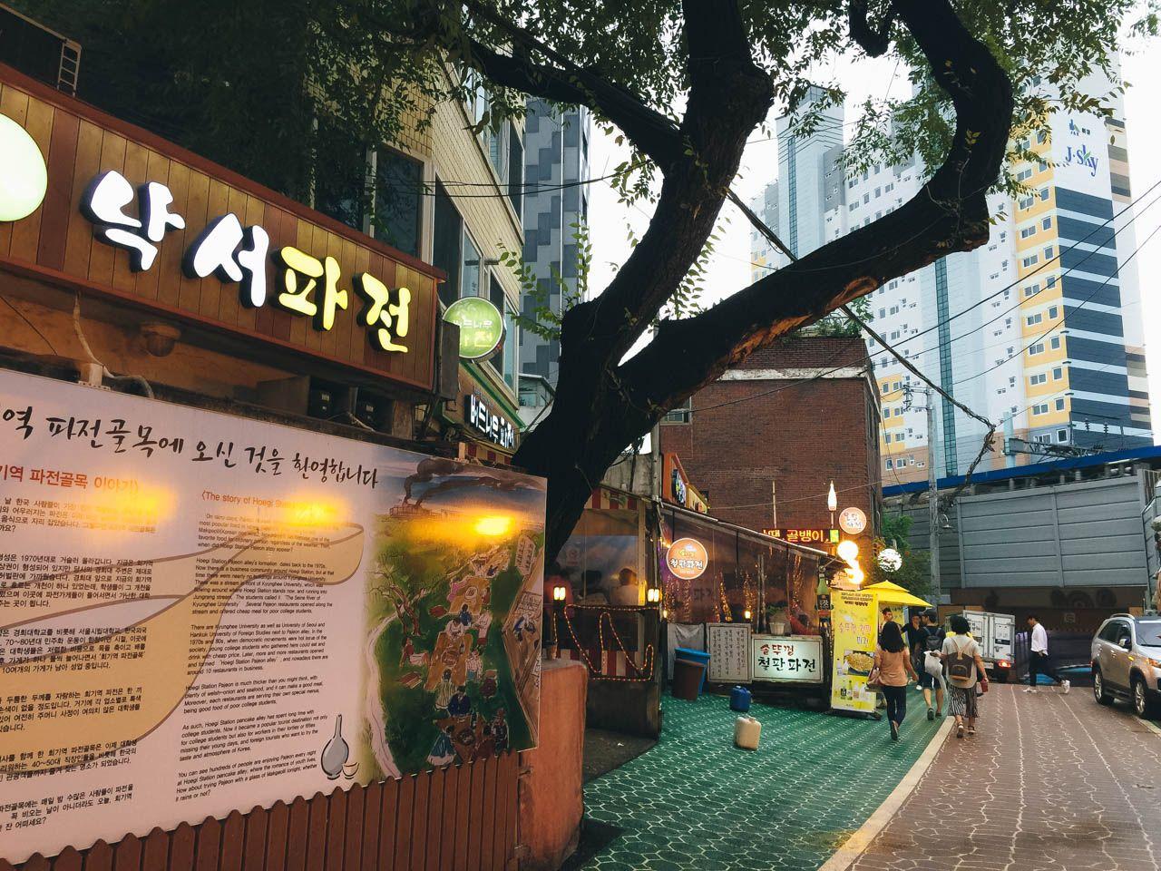 exterior view of urban building on 회기 파전골목 with trees, windows, and public space for leisure.