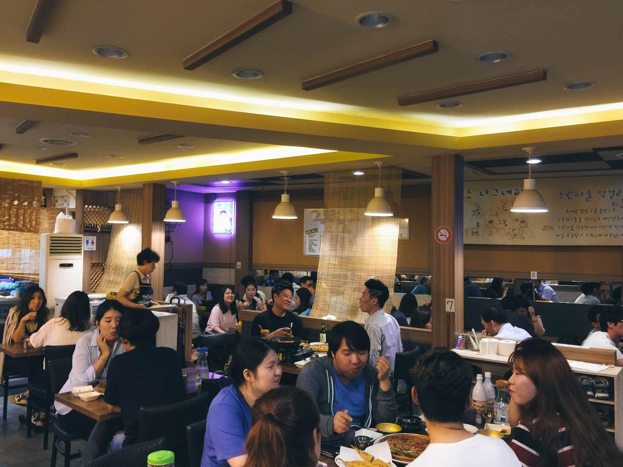 interior of restaurant in 회기 파전골목 with tables, chairs, lighting, and customers dining. Event staff in T-shirts and suits operate the building.
