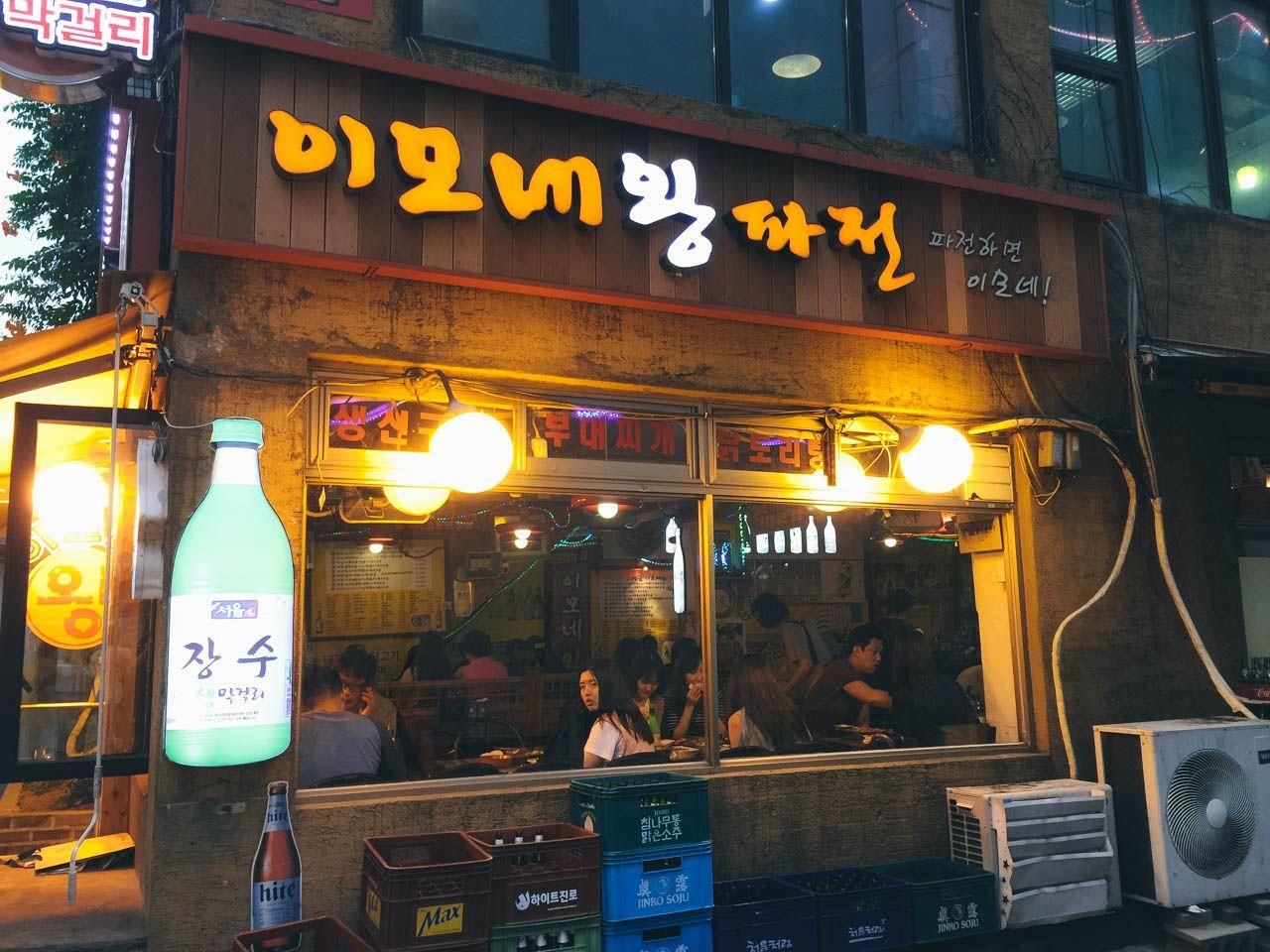 night scene of a bustling city street with a gas station, buildings, bars and restaurants, and electronic signage.