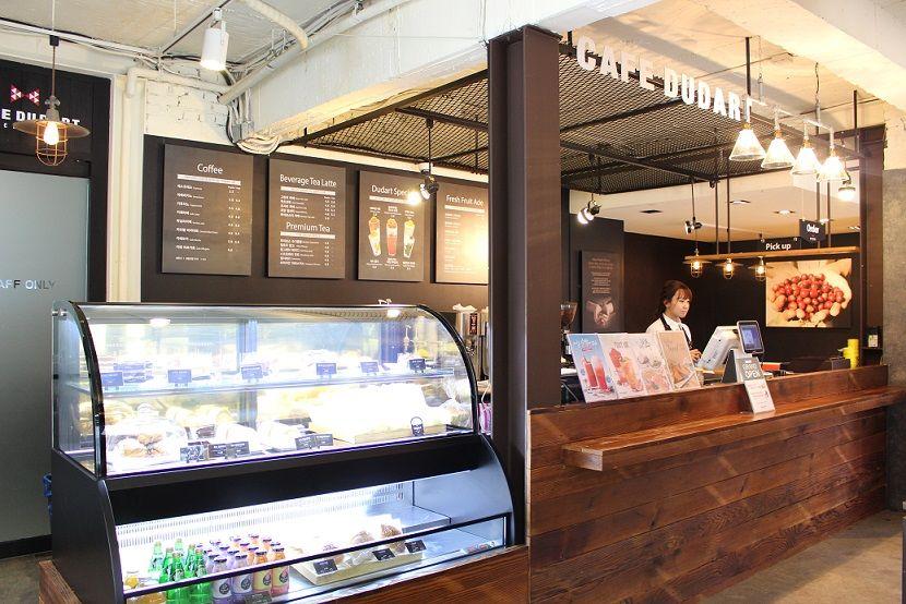 wood and glass display case showcasing delicacies in 카페두다트 with retail machines and rustic ceiling.