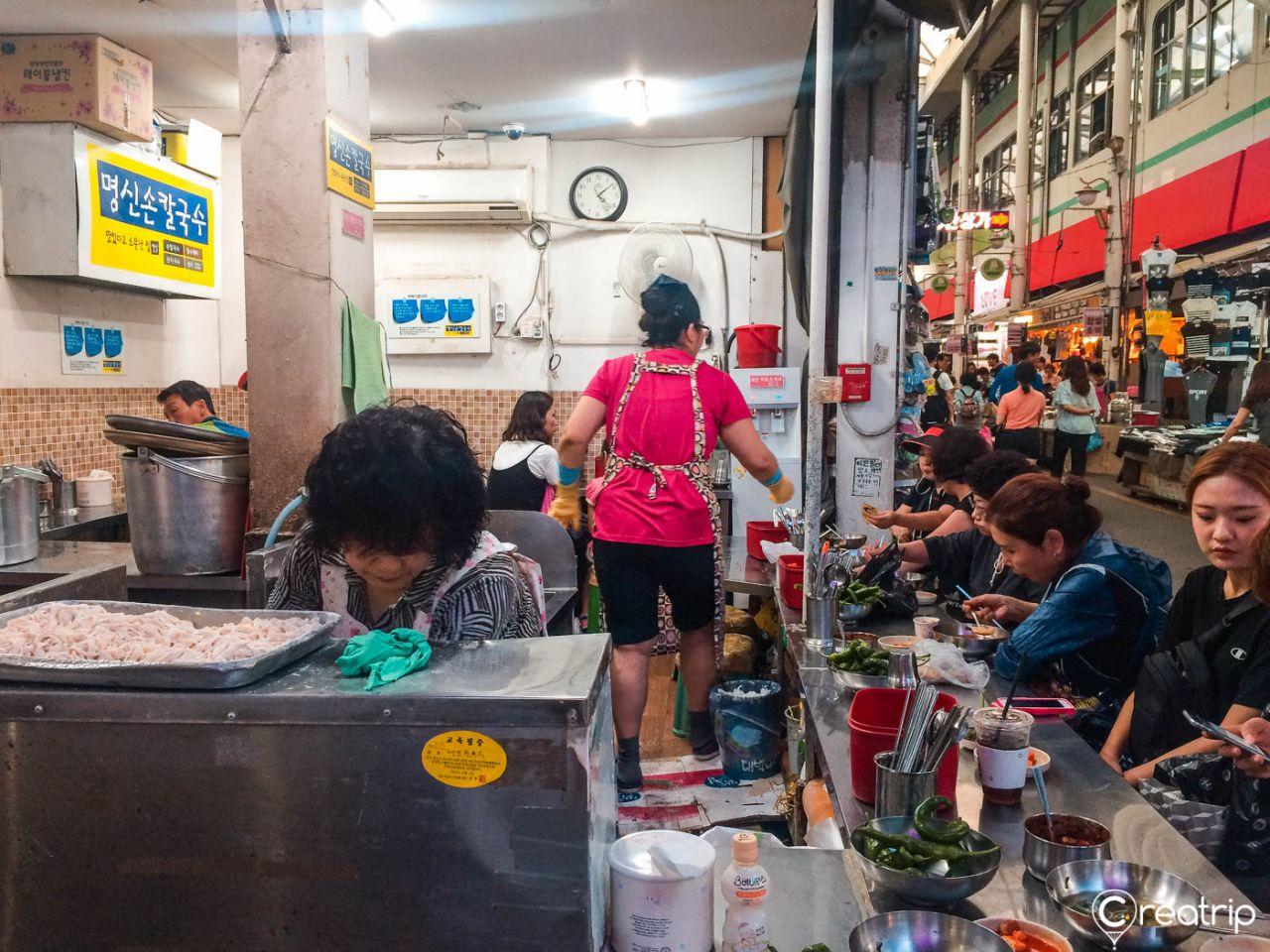 A bustling market scene in Korea's 서문시장, with a clock tower, buildings, and customers browsing trade events and clothing stalls.