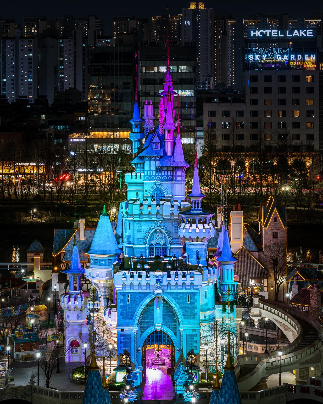 Nighttime cityscape of Seoul's Lotte World with castle-like building, skyscrapers, and landmarks visible.