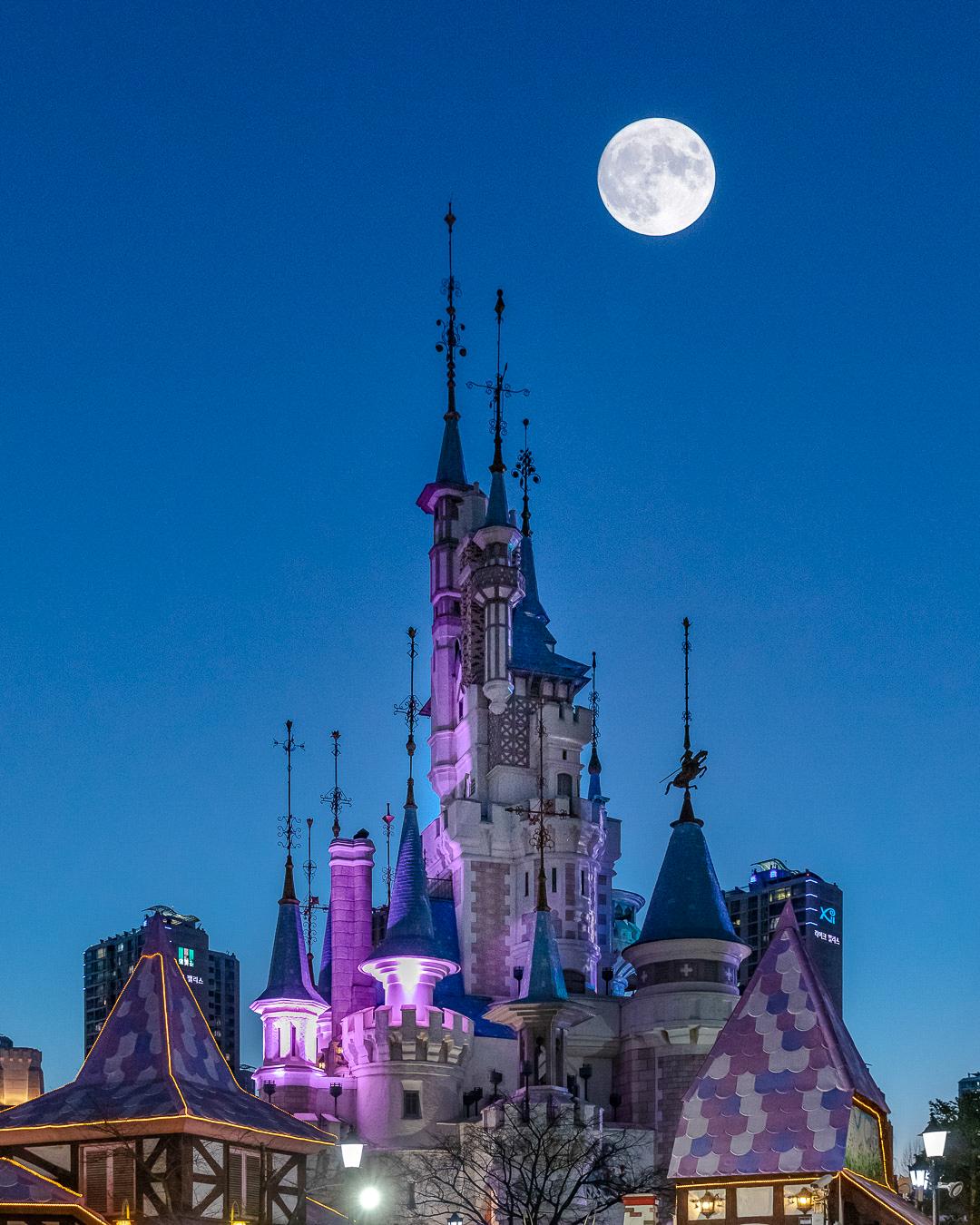 Seoul's iconic Lotte World Castle illuminated at night, surrounded by skyscrapers and a bright moon in the sky.