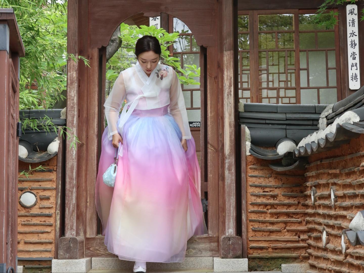 Two women wearing traditional Korean hanbok standing outside a rental shop. One is wearing a purple outerwear and the other a pink one-piece dress with plant patterns. The shop's entrance door can be seen in the background.
