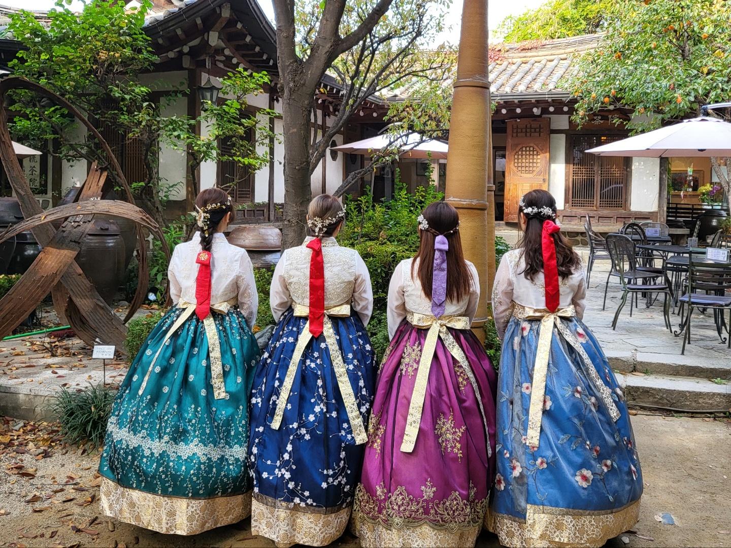 Girls having fun at Gyeongbokgung hanbok rental shop's outdoor area with plants, trees, umbrellas, tables, and entertainment.