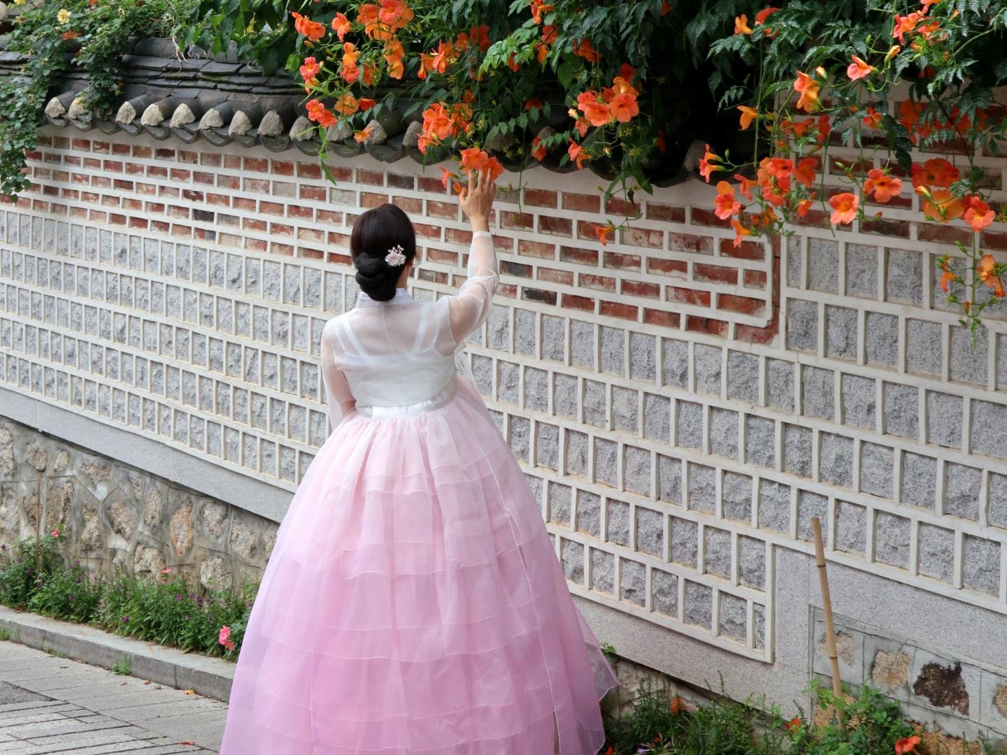 Happy woman in pink hanbok dress stands in front of Gyeongbokgung temple with beautiful plant and petal decorations.