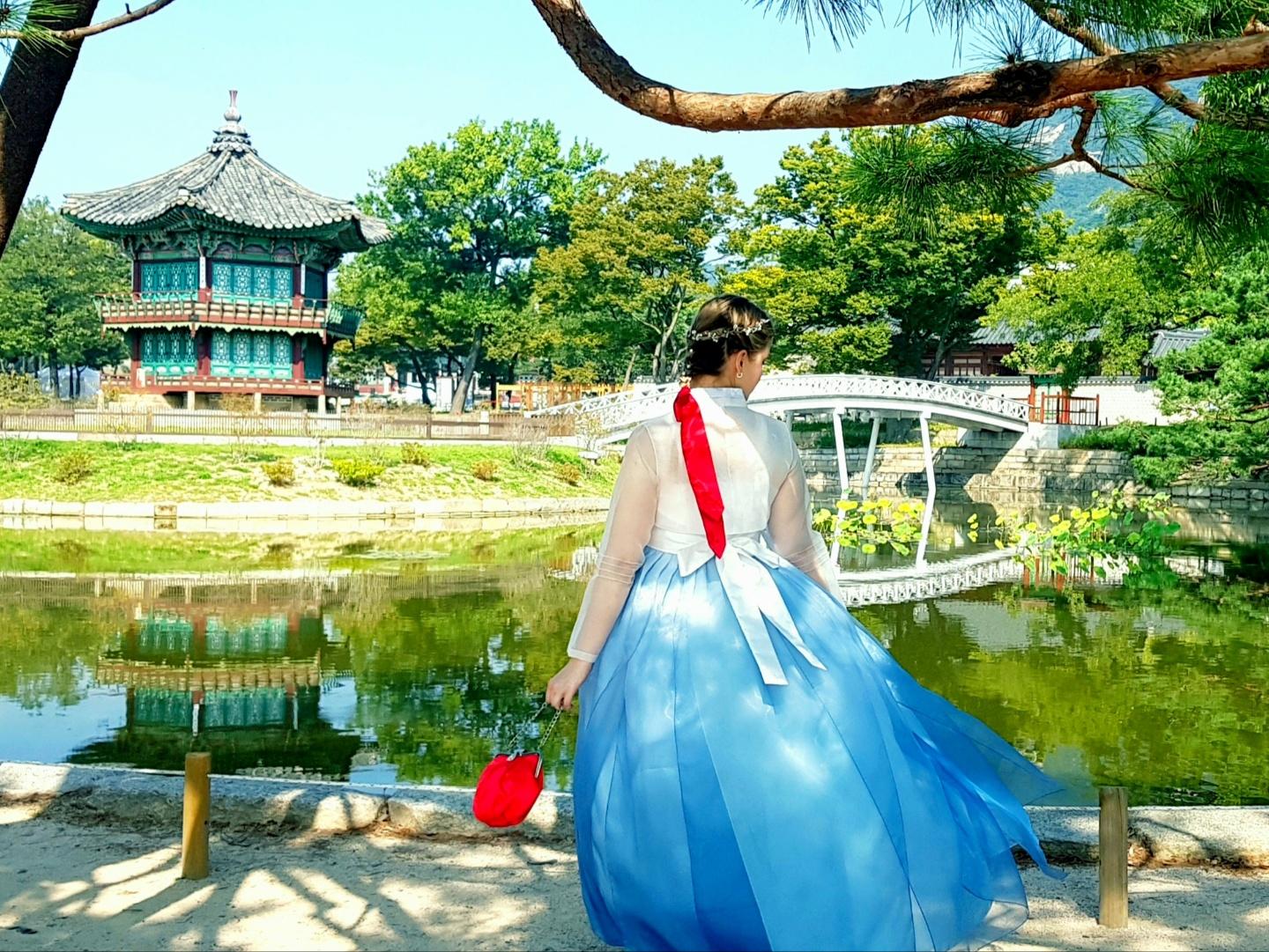 Visitors in traditional Korean dresses rent hanbok at Gyeongbokgung palace rental shop near a water feature, surrounded by lush greenery and trees with a temple in the background.