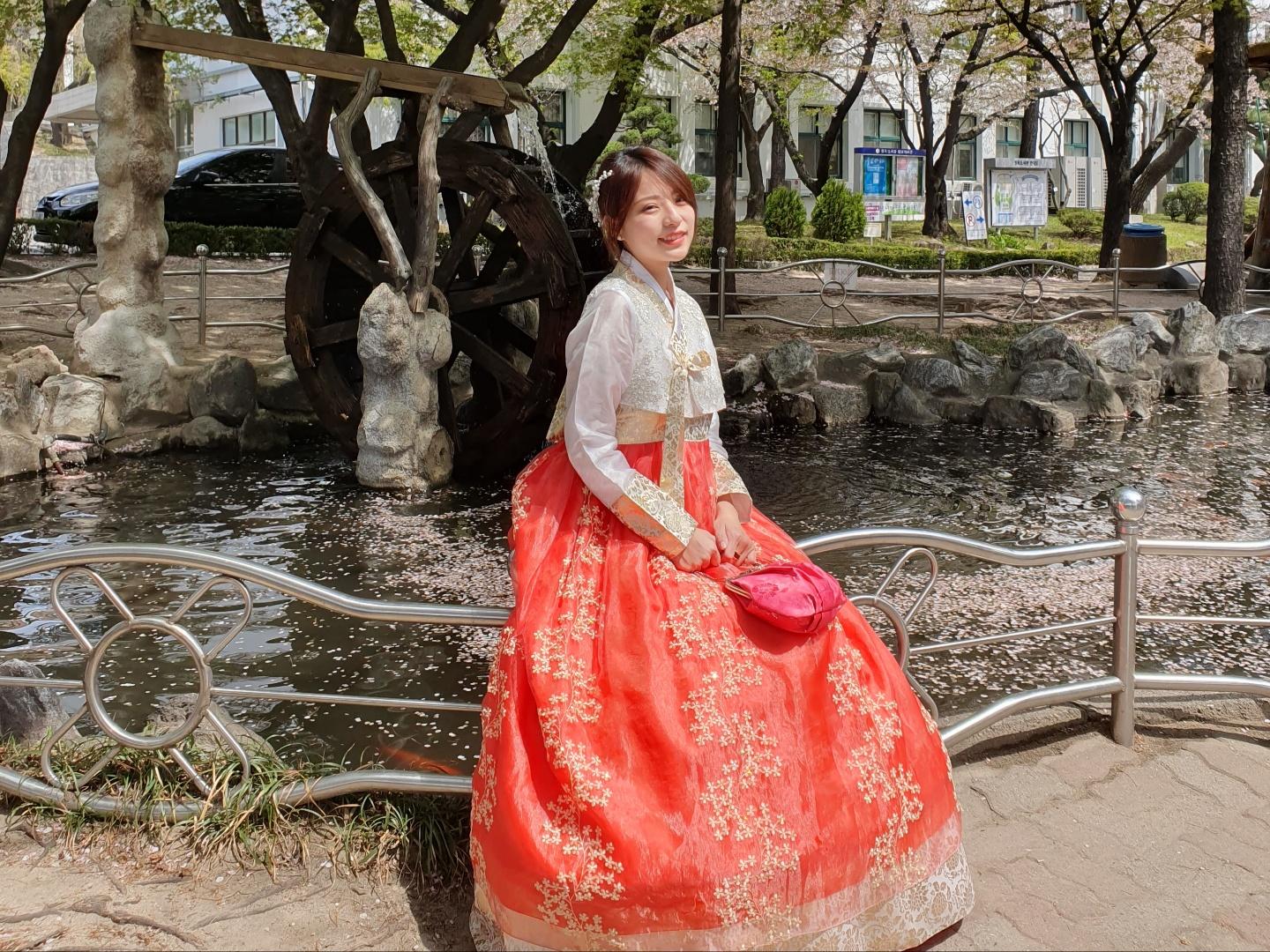 A happy woman in traditional Gyeongbokgung hanbok poses in a botanical garden near a temple, surrounded by lush trees and water.