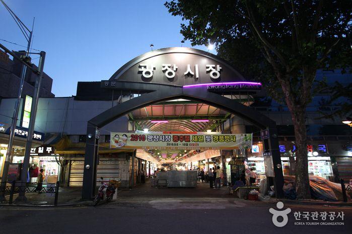 Busy entrance of the vibrant Gwangjang Market in Korea, featuring a mix of modern and traditional buildings, electronic signage, trees, and awnings.