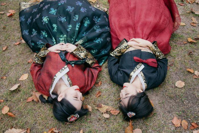 Two women laid on grass wearing traditional Korean hanbok in Gyeongbokgung, with bags and sunglasses beside them.