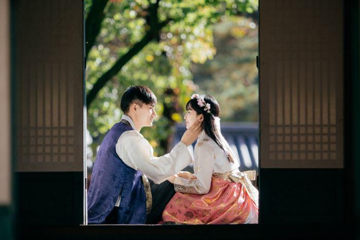 Happy couple in Gyeongbokgung hanbok rental shop dressed in formal wear sitting by tree, photographed in flash.