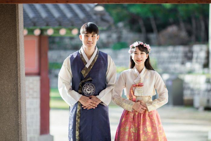 Smiling couple wearing traditional purple hanbok gown in front of Gyeongbokgung temple. Flash photography at Korean hanbok rental shop.