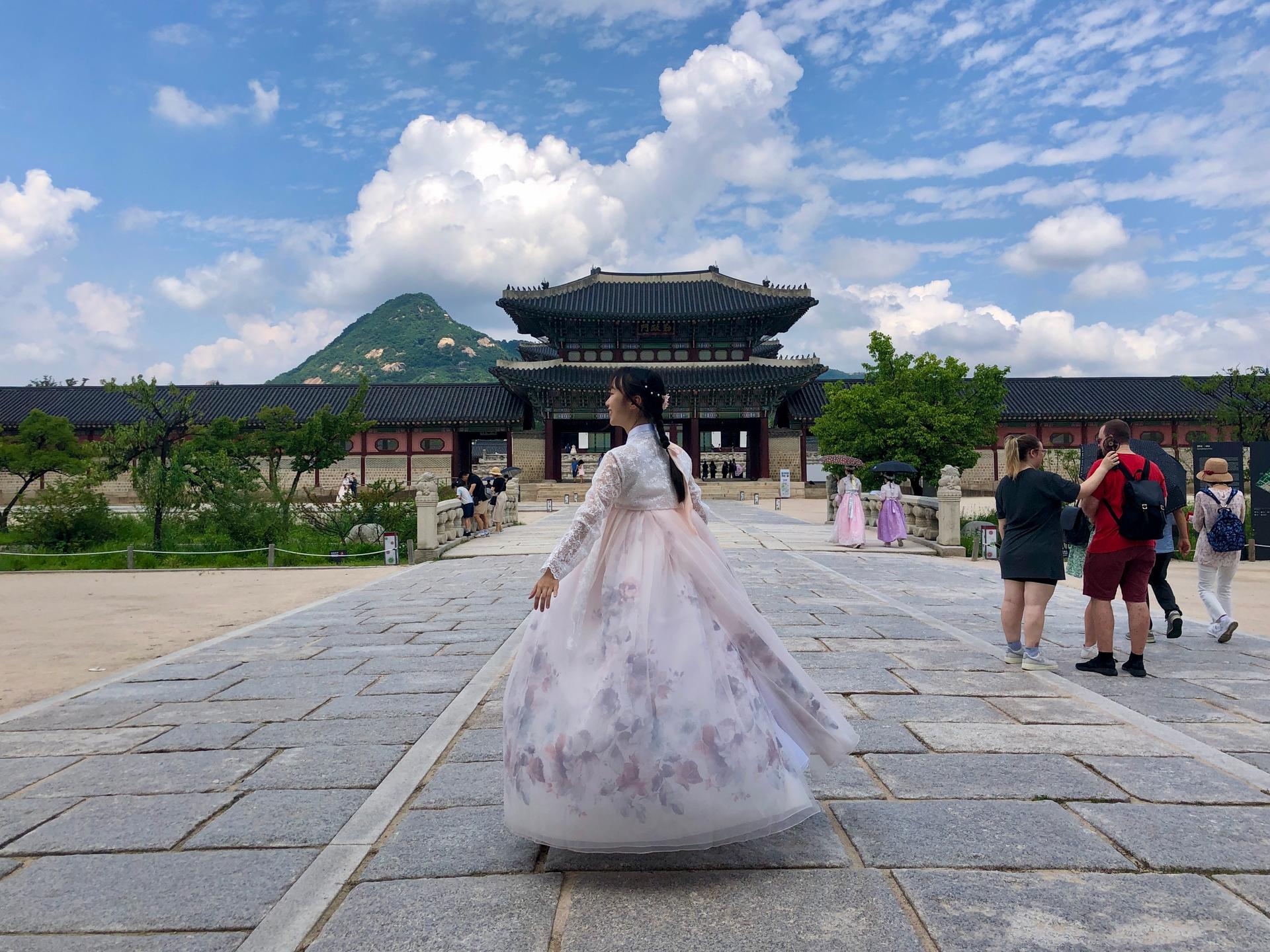 Person in hanbok dress walking towards temple against cloudy sky, surrounded by plants.