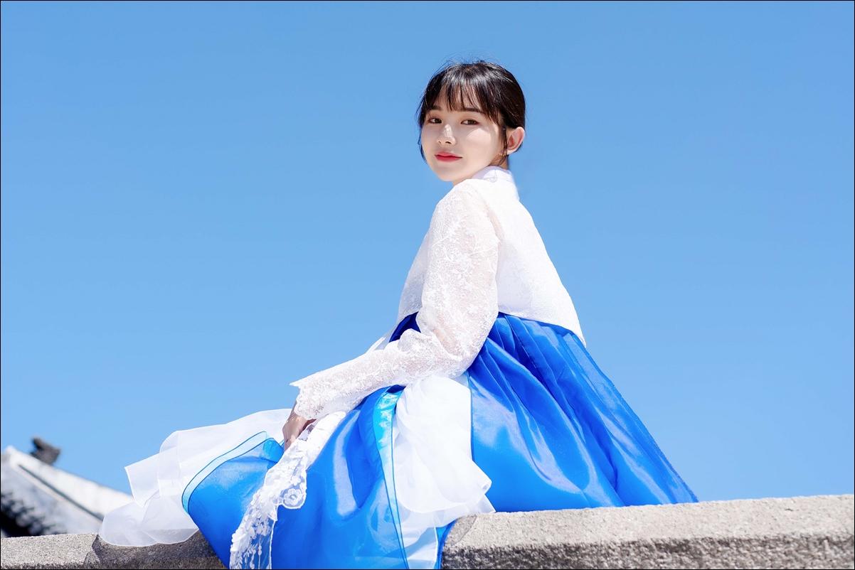 Happy woman in electric blue hanbok dress poses in front of Gyeongbokgung Hanbok rental shop, under a clear sky.