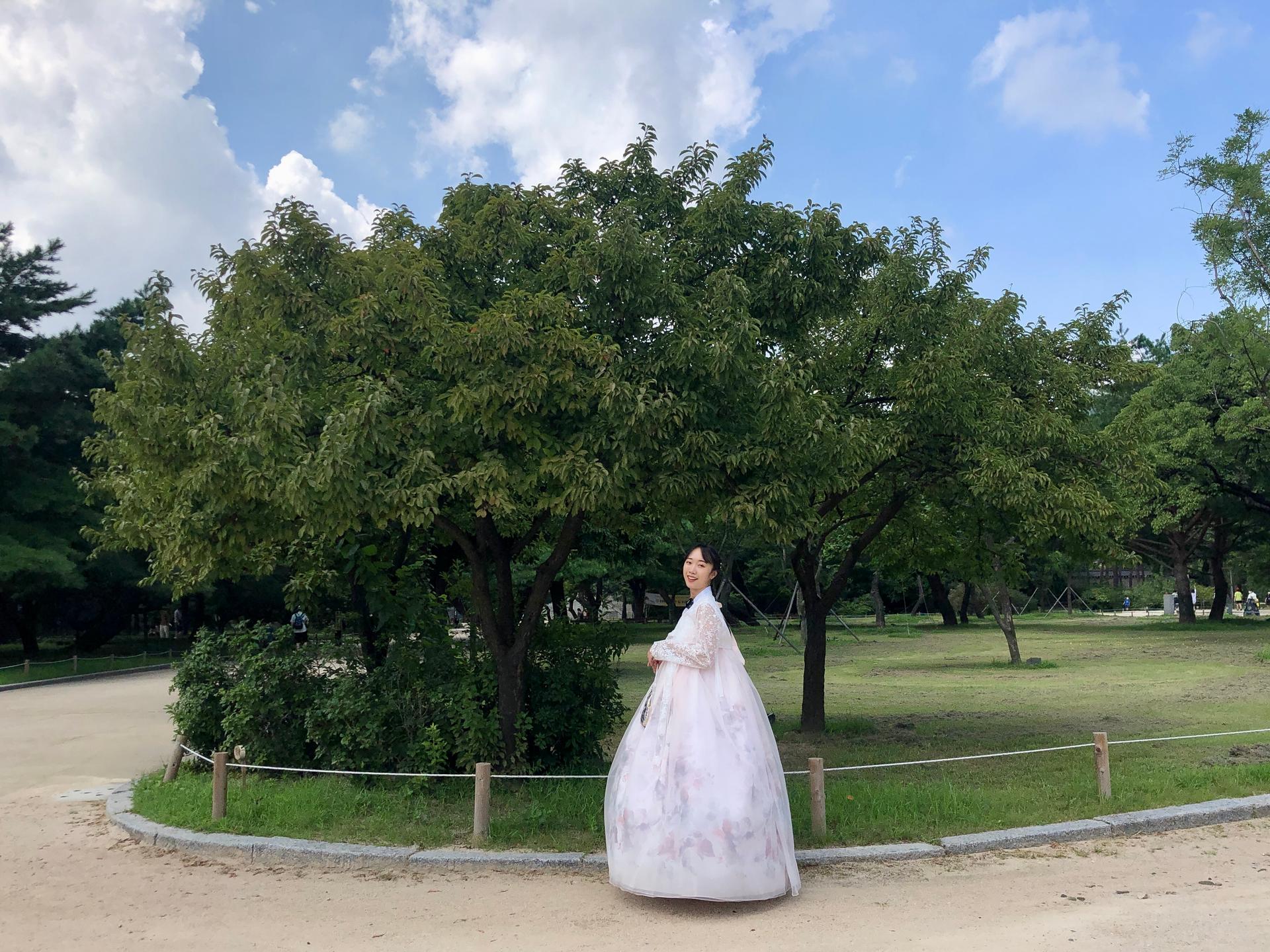 A woman wearing a hanbok stands in front of Gyeongbokgung Palace, surrounded by lush trees and a cloudy sky.