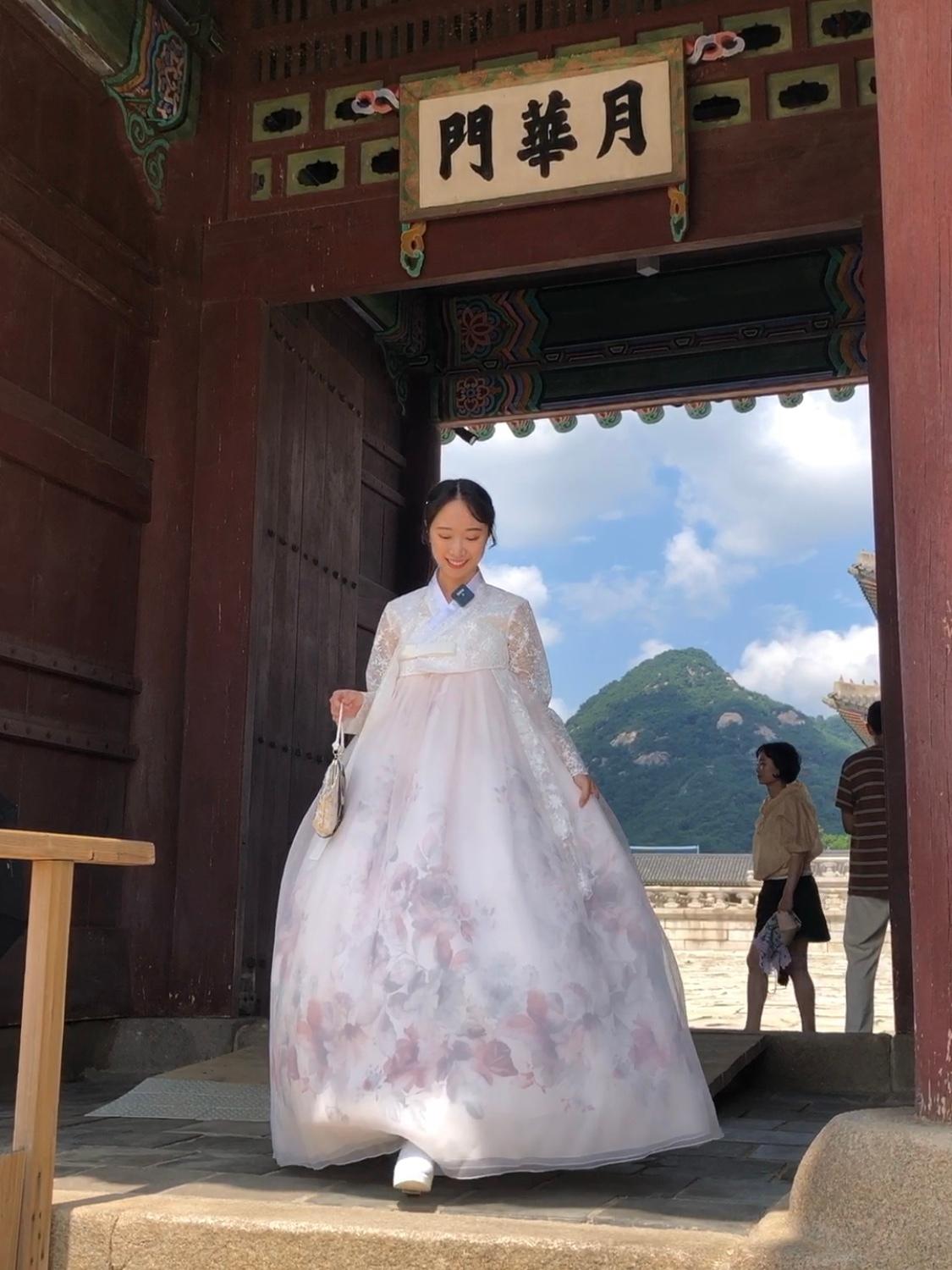A happy woman in a traditional Korean outfit stands in front of Gyeongbokgung Palace with a fox statue, temple, sky and clouds in the background.