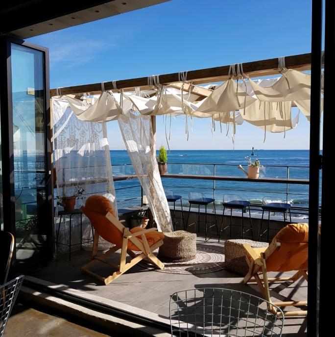 Outdoor rooftop cafe with water views, shaded chairs, wooden furniture, and sea in background.