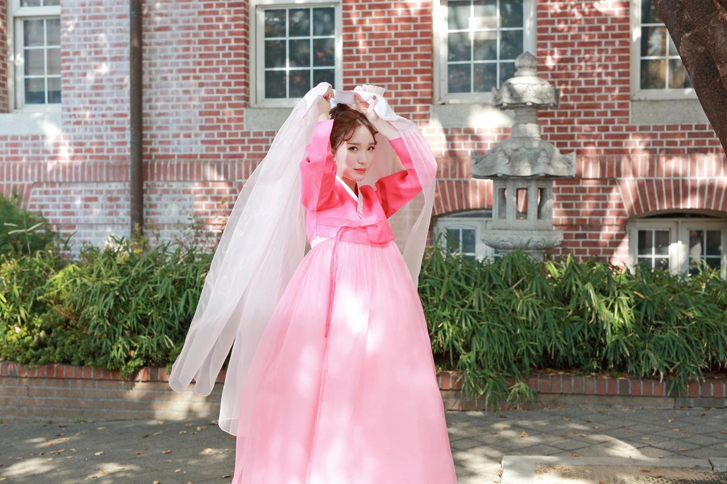 Model wearing a pink Hanbok dress standing window surrounded plants and grass.