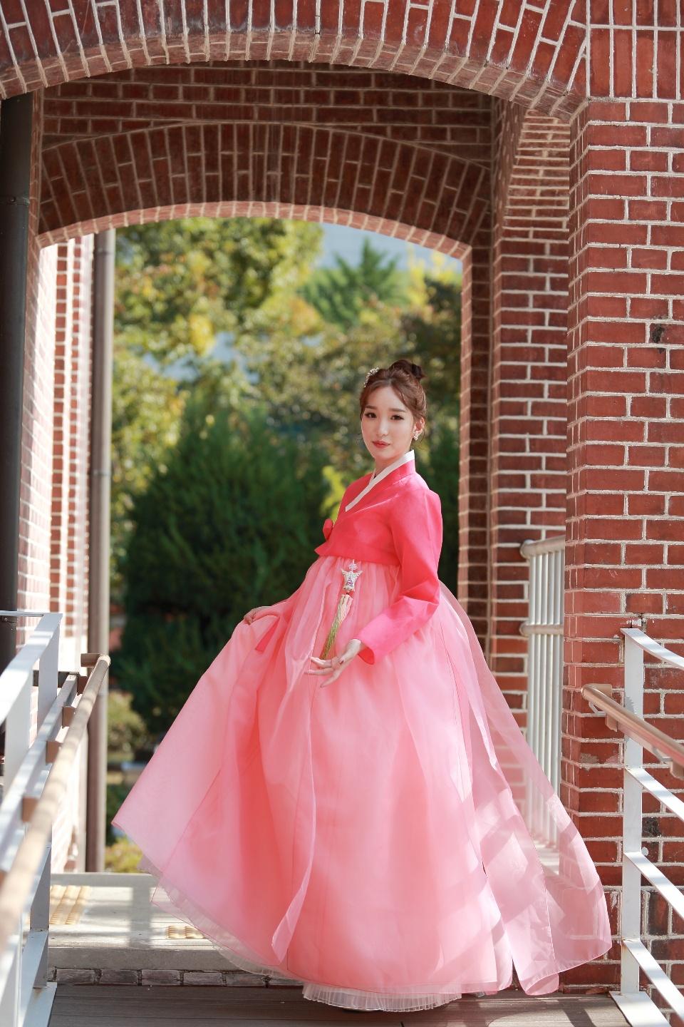 A model standing in front of <철수와영희>, a Busan Hanbok rental shop, wearing a pink traditional Korean gown.