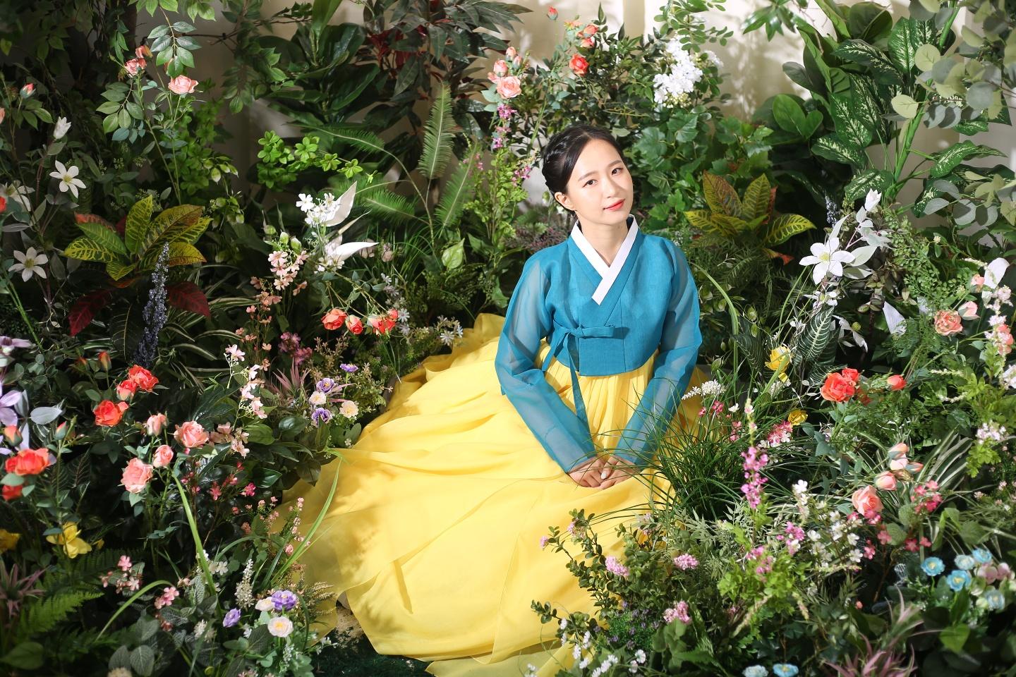 A woman wearing Hanbok stand in a lush garden surrounded by plants and flowers.