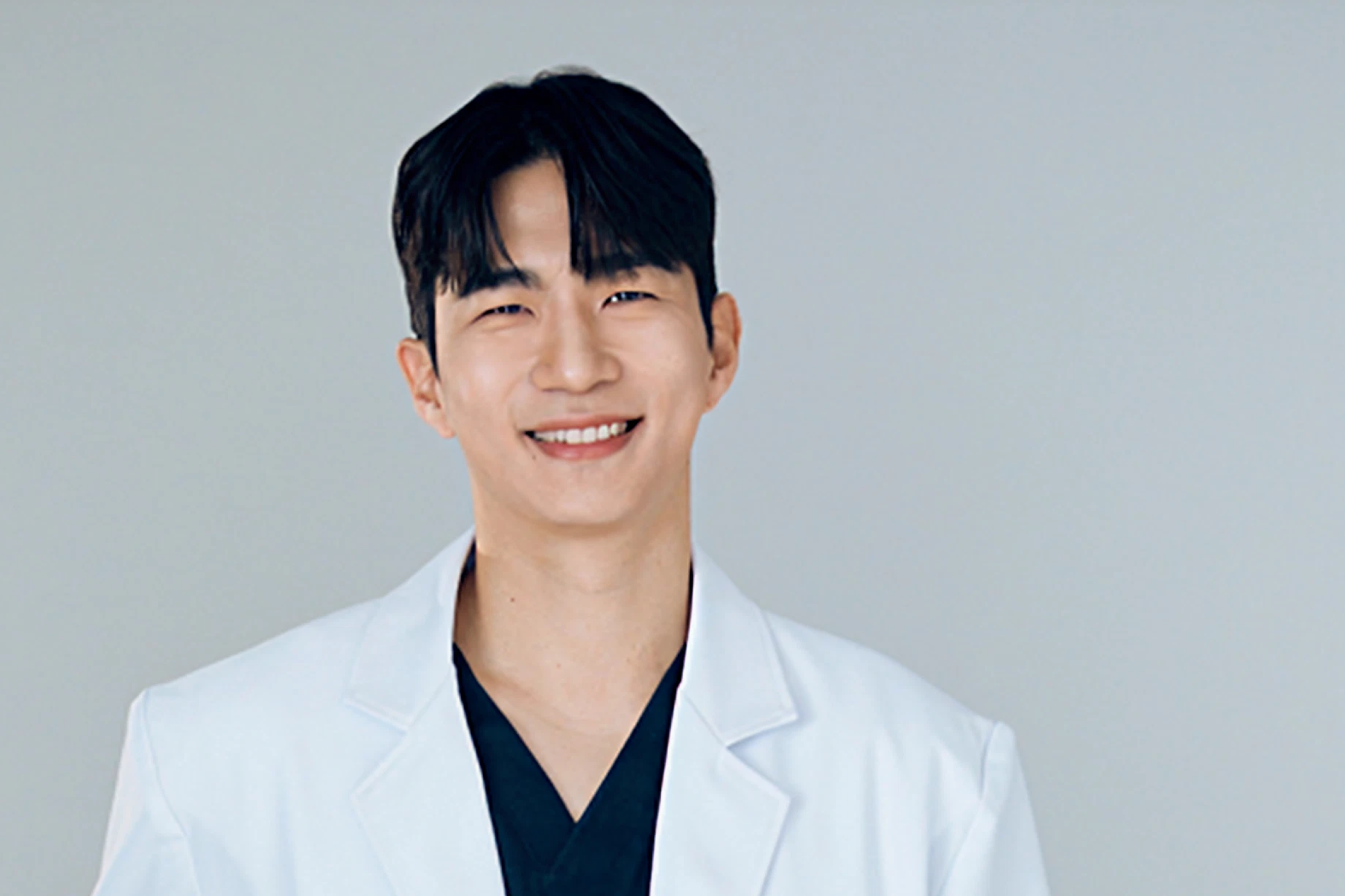 흰 가운과 검은 진료복을 착용한 피부과 전문의 상반신 사진, 창백한 배경과 프라이빗한 진료 환경을 강조하는 이미지