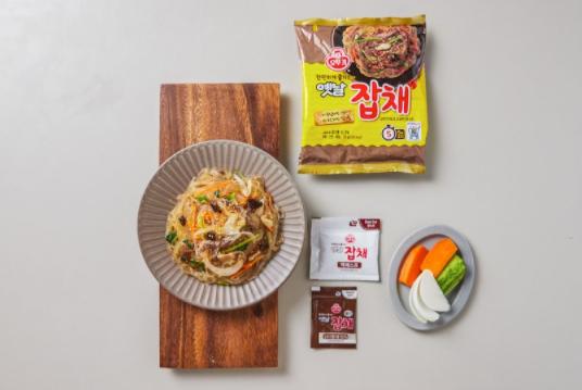 korean brand ottogi's Japchae pack next to plate of japchae on wooden platter and a plate of vegetable, and seasoning packets