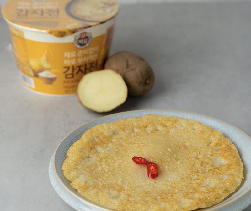 korean potato pancake on a plate next to potatoes  and korean brand beksul's potato pancake instant cup mix container 